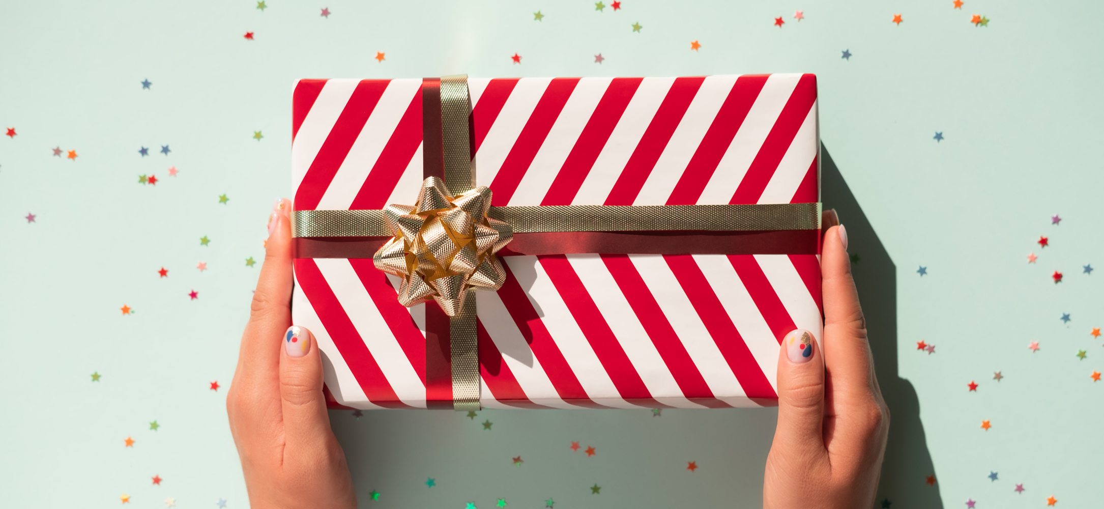 Two hands holding a rectangular holiday gift wrapped in red and white striped wrapping paper and a gold bow.